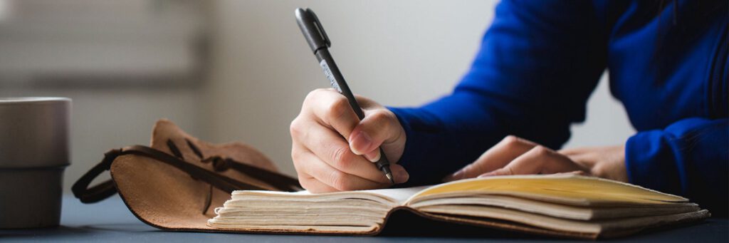 A woman's hand holds a black pen and is writing in a leather bound journal. Her face is not visible, but her torso and arm are dressed in bright blue.