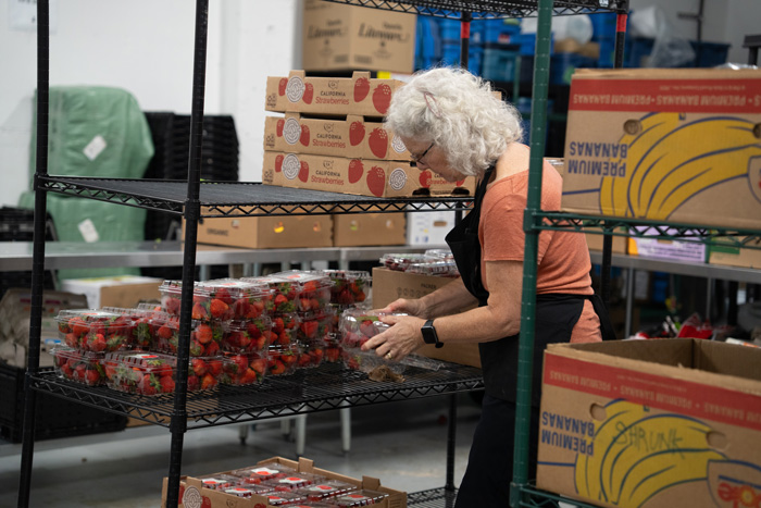 A grey-haired volunteer arranges containers of strawberries on a warehouse shelf beside stacked produce boxes.