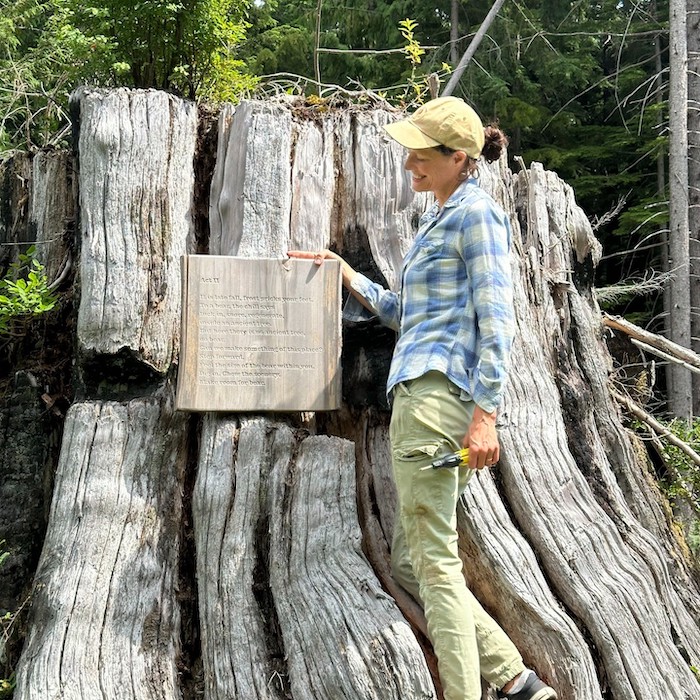 Kendra Fanconi, wearing a tan baseball cap and blue plaid shirt, stands beside a massive weathered tree stump in a forest, holding a wooden plaque with text engraved on it.